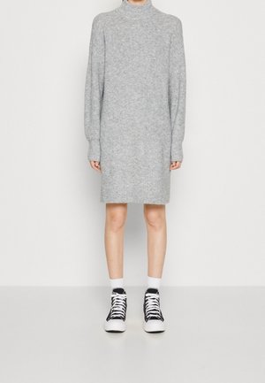 Person trägt ein graues, langärmeliges Strickkleid mit hohem Kragen sowie schwarze High-Top-Sneaker mit weißen Schnürsenkeln und weißen Socken.