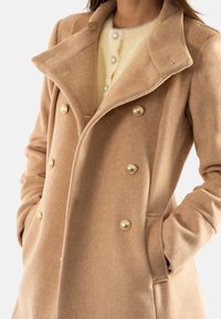 Cappotto in lana a spina di pesce beige con colletto profondo, design doppiopetto, bottoni dorati e tasche laterali.