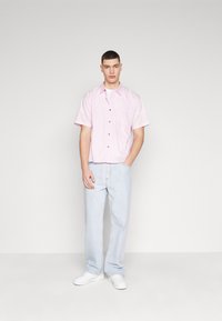 Camisa de manga corta rosa claro con botones en el frente y bolsillo en el pecho, combinada con pantalones anchos de mezclilla azul claro y zapatillas blancas.