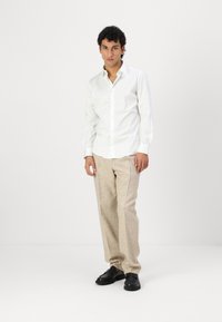 White long-sleeve shirt with a collar, paired with beige textured trousers and black shoes, standing against a plain background.