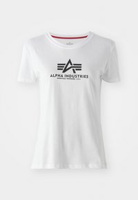 T-shirt in cotone bianco con un logo nero recante la scritta "ALPHA INDUSTRIES" e un emblema. Maniche corte, colletto a girocollo, texture liscia.