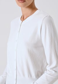 Donna che indossa un cardigan bianco semplice con bottoni e maniche lunghe, mostrata dalla zona del petto al mento su uno sfondo neutro.