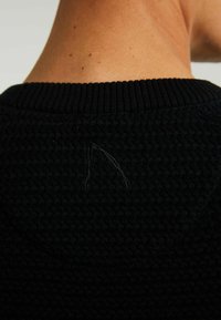 Černý pletený svetr s texturovaným vzorem a žebrovaným límcem. Významný detail: malý smyčka volné nitě na zadní straně poblíž výstřihu.