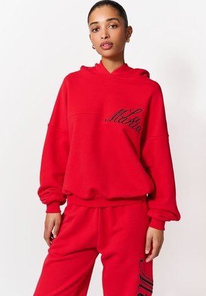 Pehmeast kangast punane kapuutsiga dressipluus, millel on avar lõige, kängurutasku ja esiküljel musta kaldkirjaga tekst. Sobivad punased püksid.