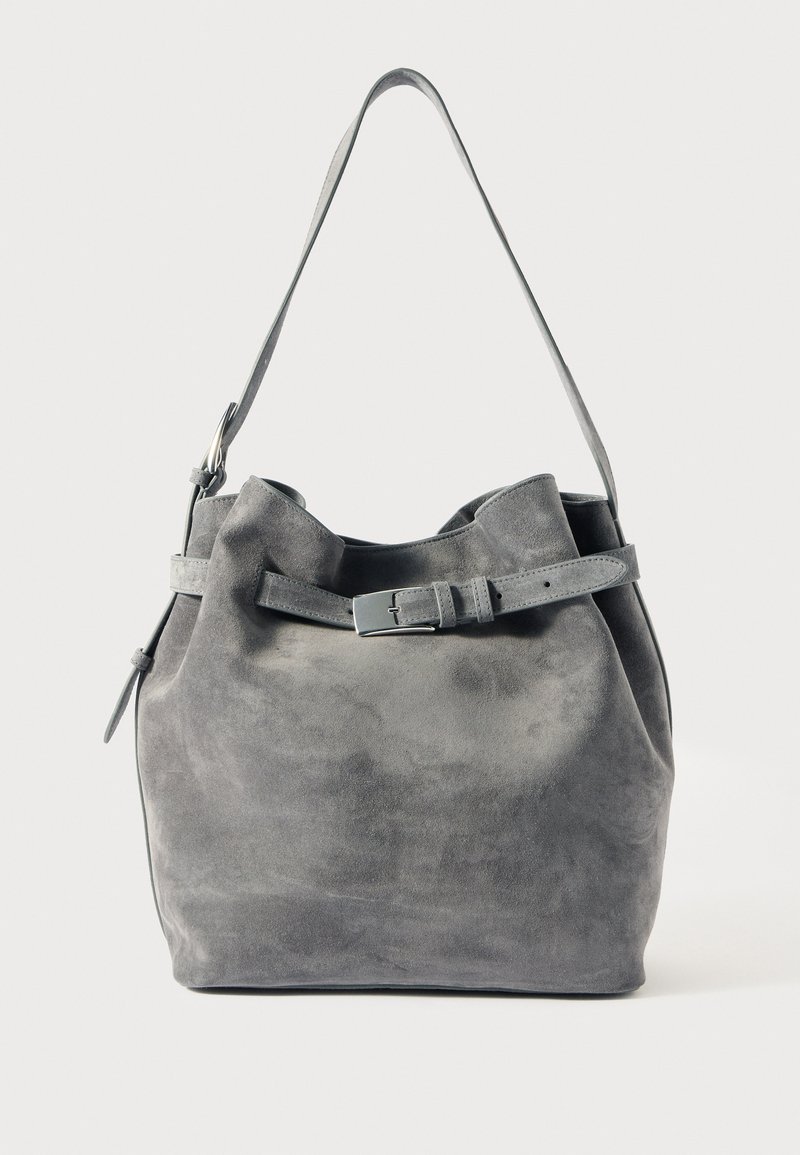 Sac seau en daim gris avec une forme structurée. Présente une large ouverture, une sangle en cuir réglable et un accent de style ceinture autour du haut.