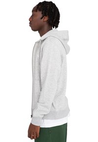 Mladý muž s krátkymi skrútenými vlasmi, oblečený v svetlo šedej zipsovej mikine, pod ňou biele tričko a tmavozelené nohavice, stojaci v bočnom výhľade.