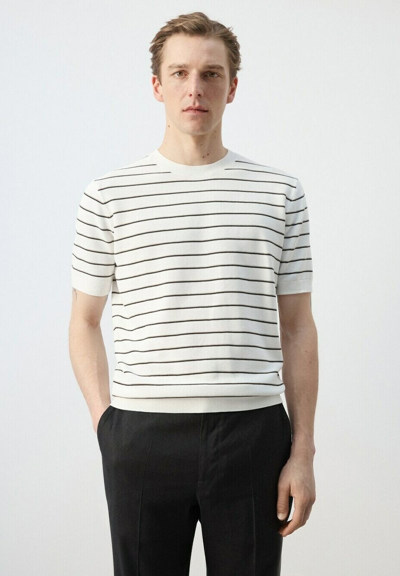 Jeune homme aux cheveux châtain clair portant une chemise blanche à manches courtes avec de fines rayures horizontales noires et un pantalon noir, debout avec une main dans la poche.