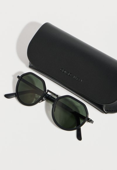 Μαύρα εξαγωνικά γυαλιά ηλίου με σκούρους πράσινους φακούς δίπλα σε μια μαύρη δερμάτινη θήκη με το λογότυπο Giorgio Armani πάνω σε λευκή επιφάνεια.