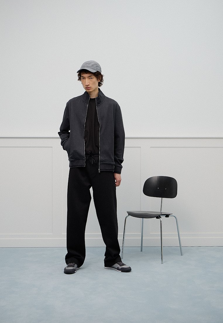 Mannenkleding met een donkere bomberjack, zwart hoodie en losse zwarte broek. Grijze pet en sneakers, met een eenvoudige zwarte stoel in de buurt.