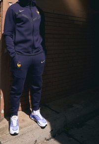 Paarse trainingsoutfit met een ritsjack en broek, voorzien van een gouden haan-embleem en het Nike-logo. De schoenen zijn wit en lichtblauw met gele accenten.