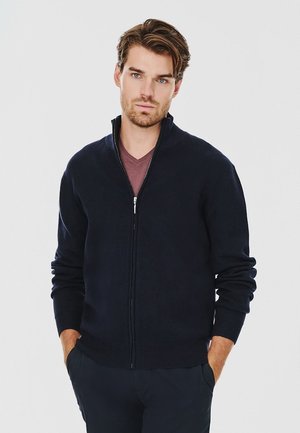 Granatowa rozpinana kardigan z prążkowanymi mankietami i dołem, wykonany z miękkiego, teksturowanego materiału, noszony na głębokiej marsala koszulce. Model pozuje z rękami w kieszeniach.