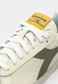 White leather sneakers with gray suede accents, textured round toe, white laces, and a yellow tongue featuring black logo and stripe.