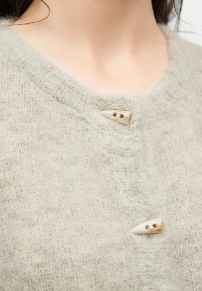 Pull doux beige duveteux avec deux petits boutons en forme de corne près de l'encolure, porté par une personne aux cheveux foncés.