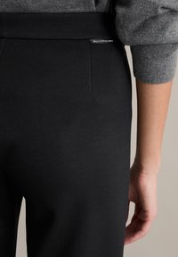 Schwarze Hose aus glattem Material mit breitem elastischem Bund und einem kleinen Markenschild auf der Rückseite. Nahaufnahme der Rückansicht mit Hand.