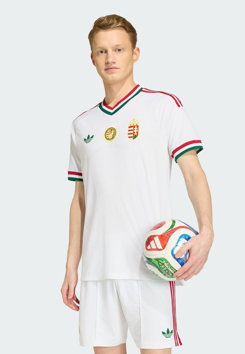 Jeune homme en uniforme de football blanc avec des bordures rouges et vertes tenant un ballon de football Adidas multicolore sur un fond uni.