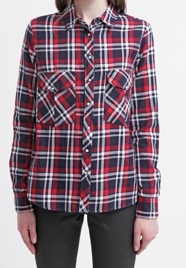 Personne portant une chemise à carreaux rouge, marine et blanche à manches longues avec deux poches poitrine et un pantalon noir.