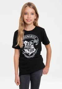 Schwarzes Baumwoll-T-Shirt mit einem Hogwarts-Wappen in Weiß, das Symbole der verschiedenen Häuser und ein Banner darunter zeigt. Mit kurzen Ärmeln.