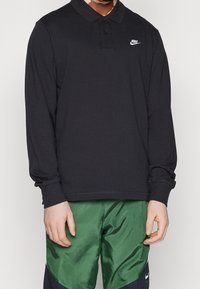 Mężczyzna w czarnym, długim rękawie, polo Nike i zielonych szortach sportowych Nike, stoi z rękami swobodnie opartymi po bokach.