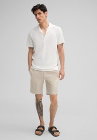 Witte polo met korte mouwen van een zachte stof, gecombineerd met lichtgestreepte beige shorts en zwarte slippers, met een zichtbare tatoeage op het been.