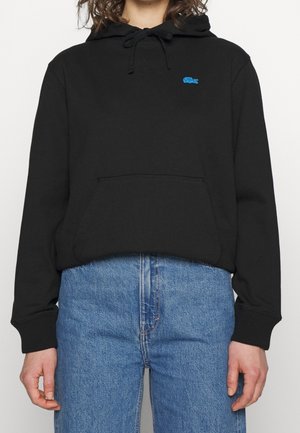 Personne portant un sweat à capuche noir avec un logo bleu sur la poitrine et un jean bleu, debout devant un fond uni.
