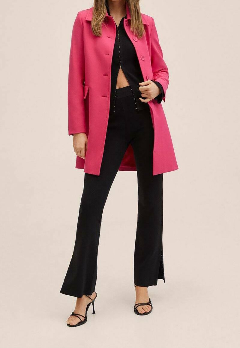 Femme portant un manteau rose vif boutonné par-dessus un crop top noir, un pantalon noir évasé avec des fentes, et des sandales à talons hauts noires avec des lanières.