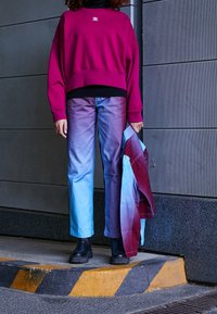 Bordowy, oversize'owy sweter z ściągaczami na mankietach, zestawiony z spodniami w gradiencie od niebieskiego do lawendowego. W rękach trzymane są czarne botki na platformie oraz wzorzysta kurtka.