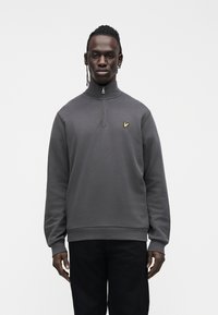 Man standing wearing a dark gray quarter-zip sweatshirt with a small yellow bird logo on the chest and black pants against a plain white background.