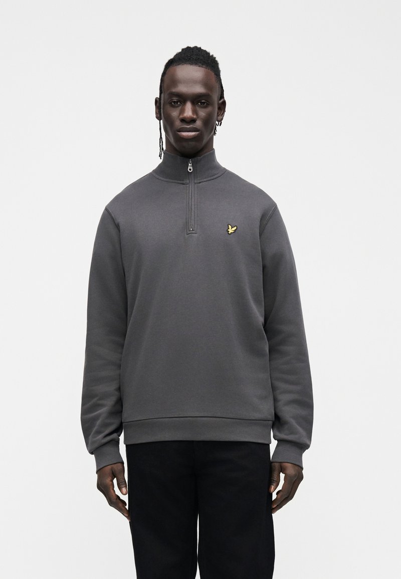 Man standing wearing a dark gray quarter-zip sweatshirt with a small yellow bird logo on the chest and black pants against a plain white background.