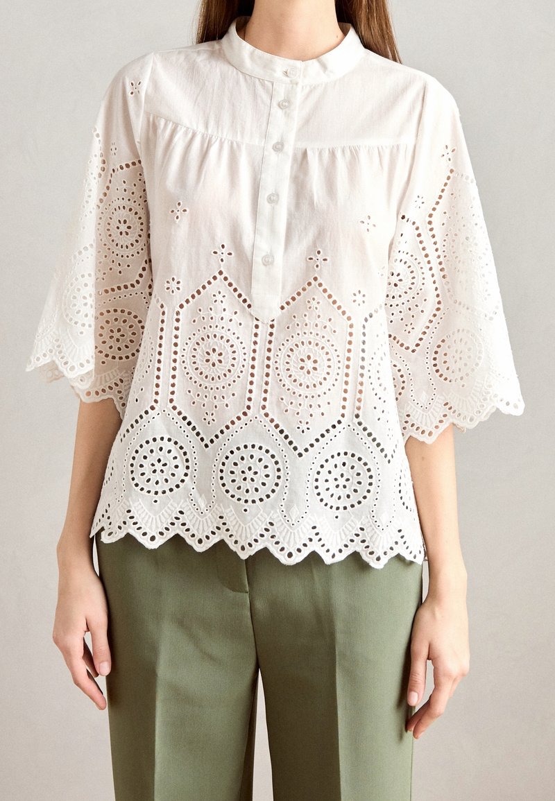 Blouse en dentelle blanche à œillets avec ourlet festonné et manches larges, portée avec un pantalon vert olive sur une personne debout devant un fond uni.