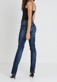 Vaqueros de mezclilla azul marino con corte ajustado, que cuentan con bolsillos traseros y detalles de costura, combinados con una blusa sin mangas negra y tacones altos.