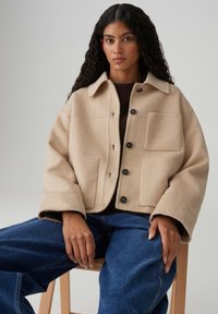 Veste en laine beige avec col, coupe oversize, grandes poches avant et boutons noirs, portée avec un pantalon en jean bleu.