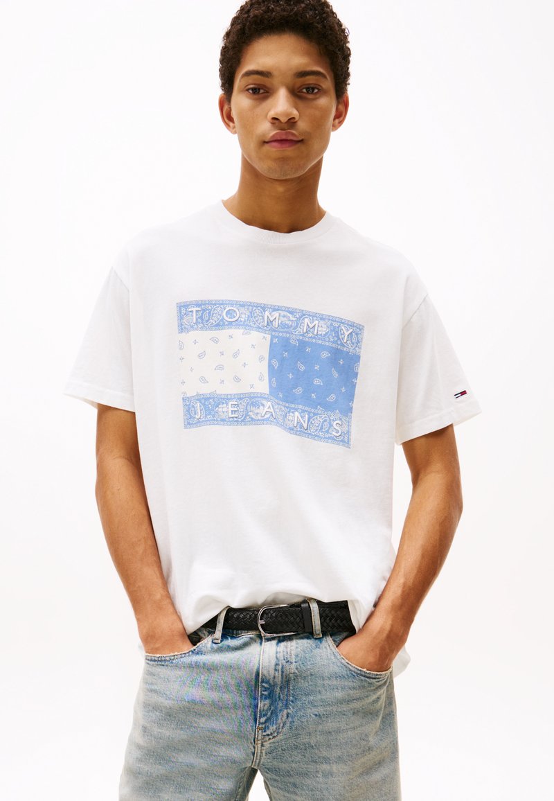 Camiseta de algodón blanca con un gráfico de patrón de bandana azul y el texto "TOMMY JEANS", combinada con jeans de mezclilla claros y un cinturón trenzado negro.