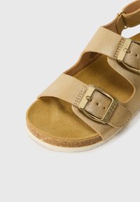 Lähivõte beežist nobedast sandaalist, millel on korktalda ja kaks reguleeritavat rihma, mis on kinnitatud kuldsete metallist pandlitega valgel taustal.