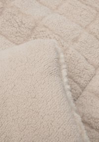 Gestructureerde beige deken van pluizige stof, met een vierkant patroon met subtiele variaties in textuur en een zachte, vlezige rand.