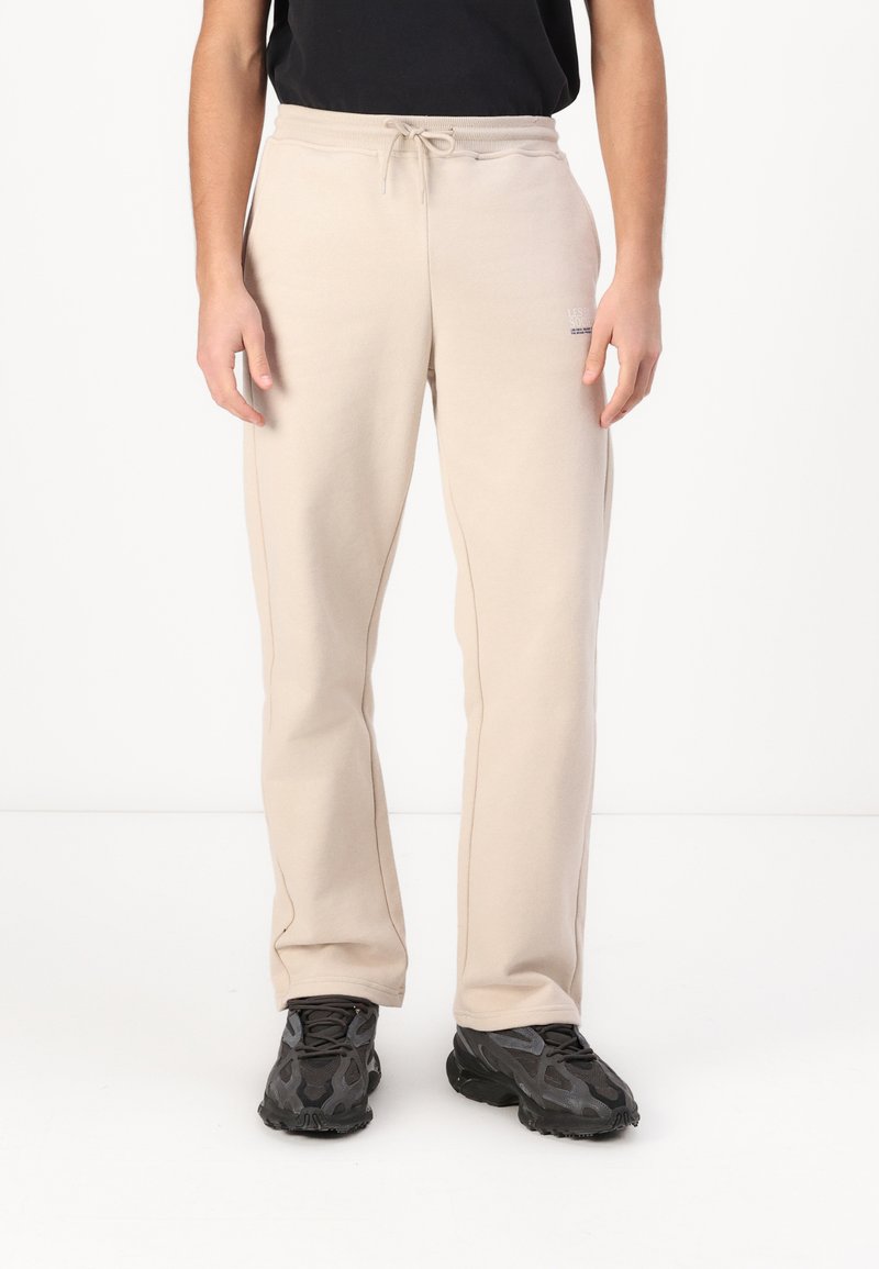 Beige Jogginghose mit Kordelzugbund, seitlichen Taschen und einer sauberen Passform. Getragen mit schwarzen strukturierten Sneakern.