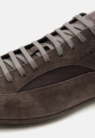 Καφέ δερμάτινο αθλητικό παπούτσι από σουέτ με γκρι κορδόνια και πλέγμα πλαϊνή επιφάνεια, που αποκαλύπτει λεπτομερείς ραφές και υφή σε λευκό φόντο.