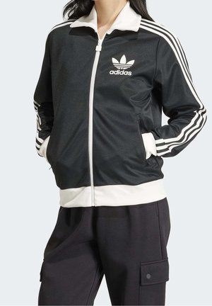 Femme portant une veste de survêtement Adidas noire et blanche avec trois bandes blanches sur les manches et le logo Adidas sur la poitrine, mains dans les poches.