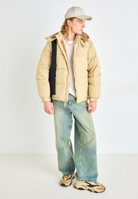 Beige puffer jacket with a hood, worn over a light textured sweater. Loose blue jeans and beige-and-black sneakers, accessorized with a cap.