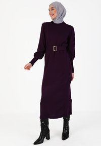 Lila langärmliges Kleid mit hohem Kragen, tailliert mit einer goldenen Gürtelschnalle, getragen mit schwarzen kniehohen Stiefeln.