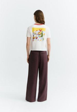 Femme aux longs cheveux bruns portant un t-shirt blanc avec des illustrations colorées de nourriture dans le dos et un pantalon large foncé, vue de dos.