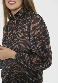 Blusa negra transparente con un cuello alto, que presenta un patrón abstracto en rosa y marrón, puños elásticos y un detalle de escote tipo ojal.