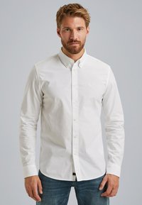 Camisa blanca de botones hecha de algodón, con cuello, mangas largas y un logotipo sutil en el pecho, combinada con jeans azules.