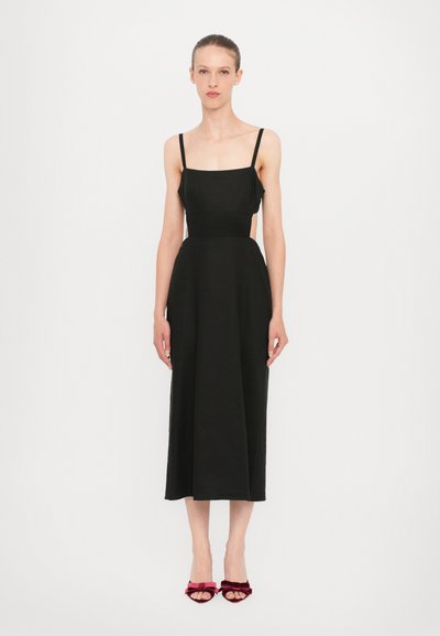 Zwarte midi-jurk met dunne bandjes en een getailleerde lijfje, met een uitlopende rok. Het model draagt rode fluwelen sandaaltjes met hakken.