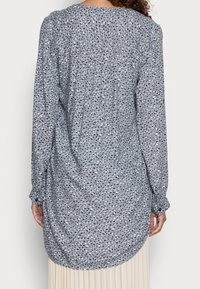 Chemise à manches longues en tissu bleu clair avec un motif éparpillé de formes bleu foncé et blanches, présentant un ourlet arrondi et des poignets à boutons.