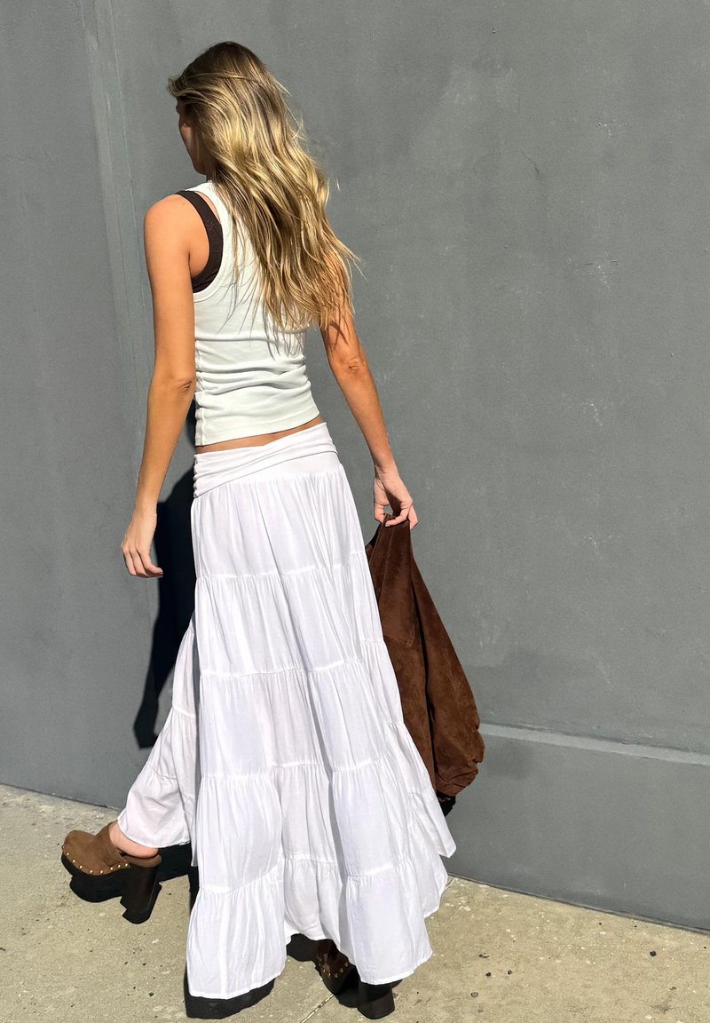 Femme aux longs cheveux blonds marche près d’un mur gris, portant une jupe maxi blanche à volants, un haut blanc sans manches, des chaussures à plateforme marron et tenant une veste marron.