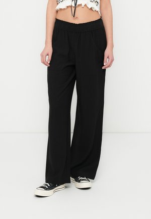 Persoană purtând pantaloni negri largi cu talie elastică, asortați cu adidași negri și albi, stând în fața unui fundal simplu și deschis la culoare.