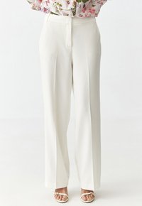 Weite Hosen in Weiß mit einem maßgeschneiderten Design, das über Faltendetails und einen flachen Bund verfügt. Getragen mit leichten, Riemchensandalen.