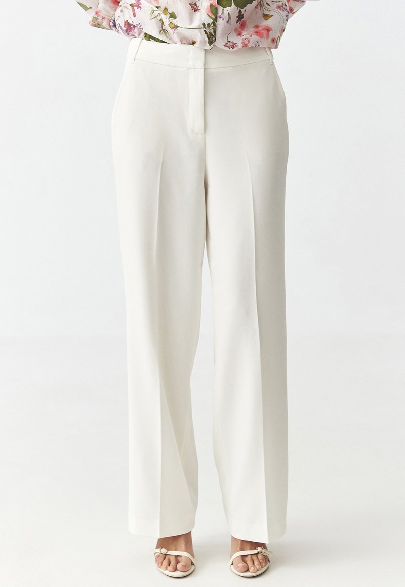 Weite Hosen in Weiß mit einem maßgeschneiderten Design, das über Faltendetails und einen flachen Bund verfügt. Getragen mit leichten, Riemchensandalen.