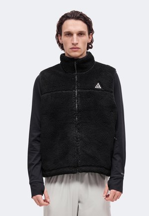 Un homme porte un gilet polaire noir texturé à fermeture éclair par-dessus un haut noir à manches longues, avec un pantalon gris clair, sur un fond uni.