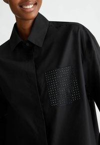 Camisa negra con collar tipo abotonado, que presenta un bolsillo texturizado adornado con pequeños studs, añadiendo contraste a la tela suave.